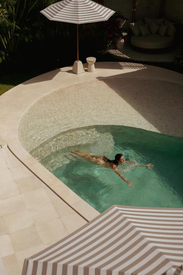 14 UNIQUE Boutique Hotels in Canggu, Bali 38 A woman swims alone in a curved, modern pool surrounded by cream-colored tiles at one of the boutique hotels in Canggu. Two striped umbrellas provide shade nearby, while lush green plants decorate the background.