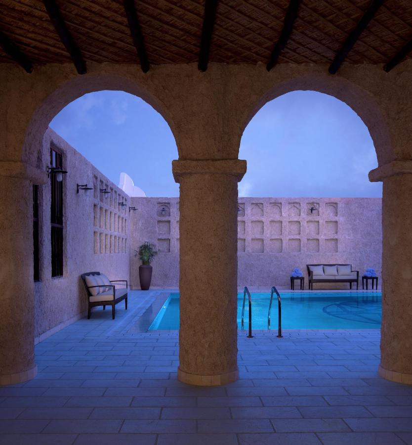 Where to Stay in Doha, Qatar: Transit, City & Beach Hotels 37 A serene courtyard with a stone archway overlooks a small, rectangular pool—ideal inspiration when searching for where to stay in Doha. Lounge chairs and a potted plant sit against textured walls, illuminated by soft evening light.