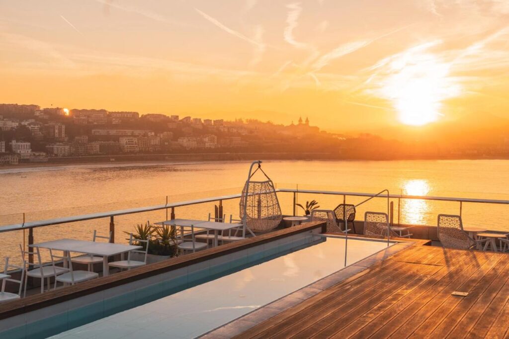 Best Boutique Hotels in San Sebastian 39 A rooftop terrace at one of the boutique hotels in San Sebastian features a pool, white tables, chairs, and a hanging seat overlooking the river and city skyline at sunrise, bathed in warm sunlight.