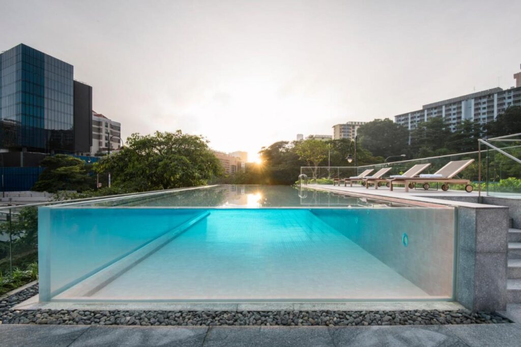 13 Most Unique Hotels in Singapore for an Incredible Holiday 18 An infinity pool with a transparent side wall, surrounded by lounge chairs and greenery, overlooks a cityscape at sunrise or sunset—one of the unique hotels in Singapore, with modern buildings in the background.