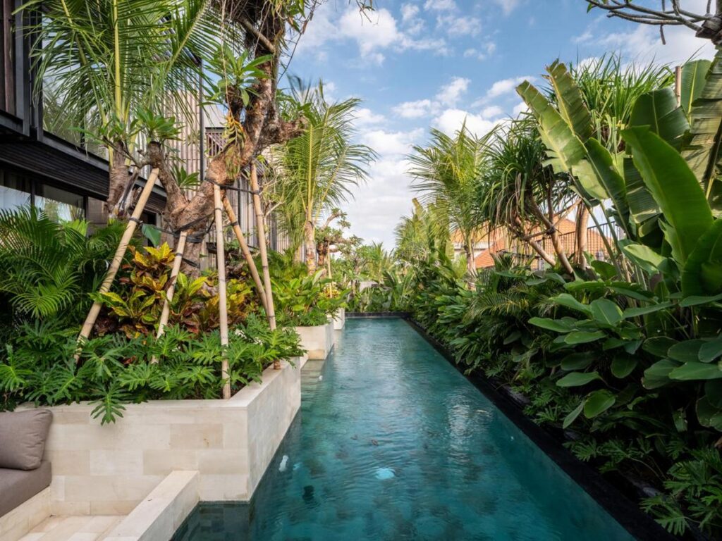 14 UNIQUE Boutique Hotels in Canggu, Bali 53 A narrow swimming pool surrounded by lush tropical plants and palm trees, with clear blue water and a partly cloudy sky above—an oasis often found at boutique hotels in Canggu, bordered by sleek modern buildings.