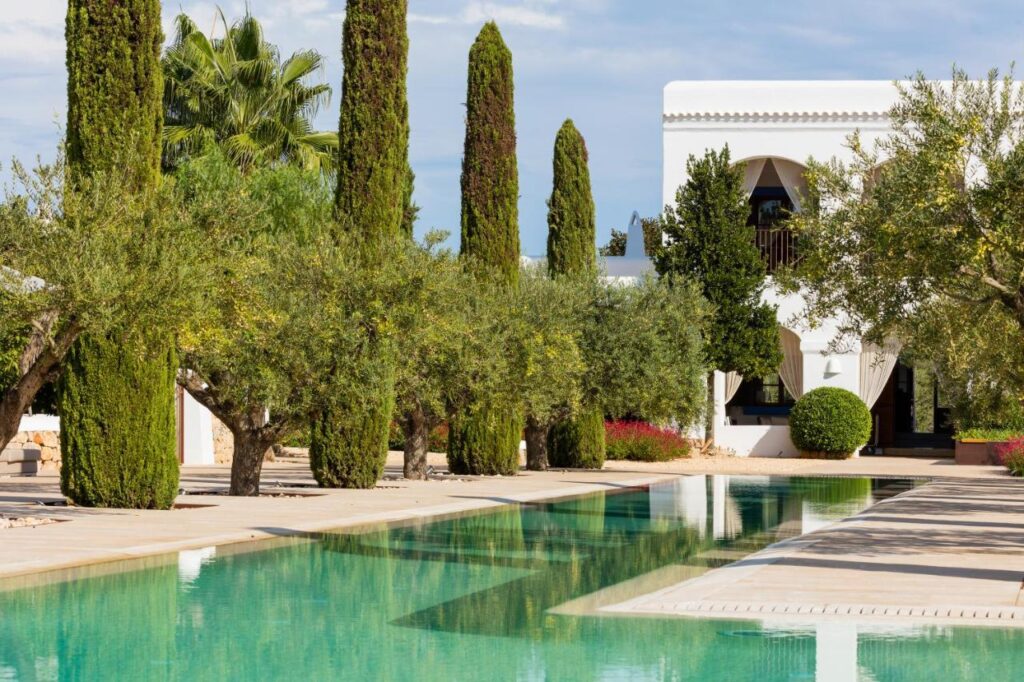 10 AUTHENTIC Finca Hotels in Ibiza 37 A modern white villa with arched windows, reminiscent of finca hotels in Ibiza, is surrounded by tall cypress trees and lush greenery, featuring a rectangular outdoor swimming pool reflecting the landscape.