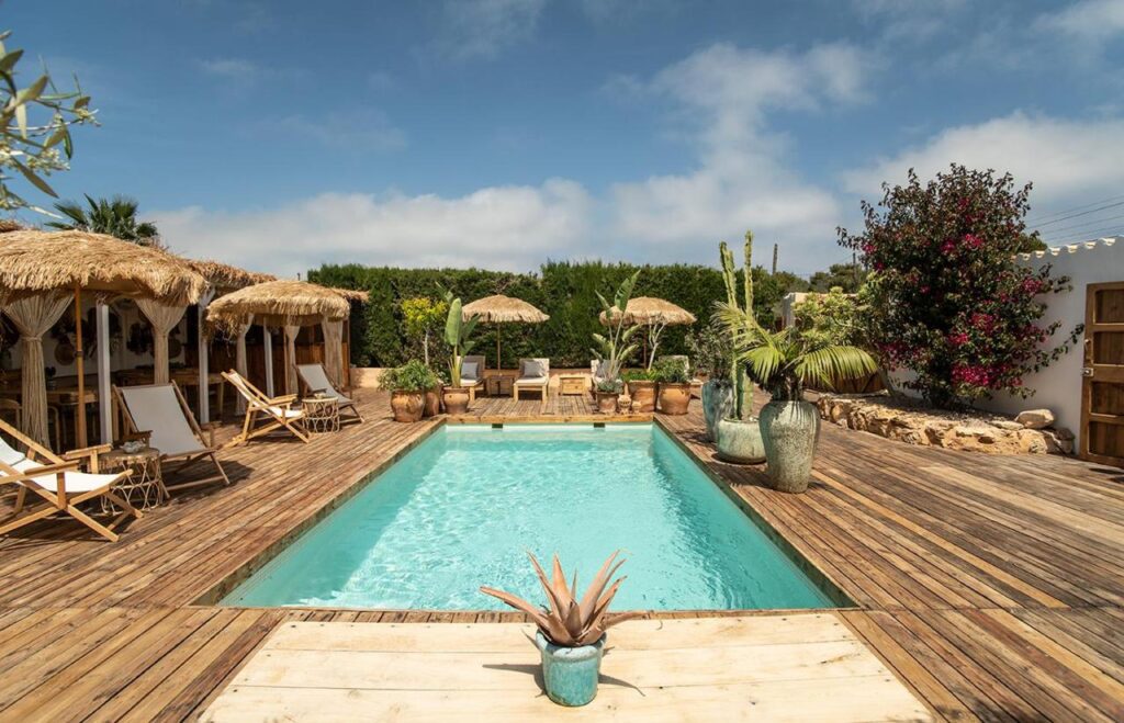 A rectangular swimming pool surrounded by wooden decking, with straw-roofed cabanas, lounge chairs, and tropical plants evokes the serene ambiance found at boutique hotels in Formentera under a clear blue sky.