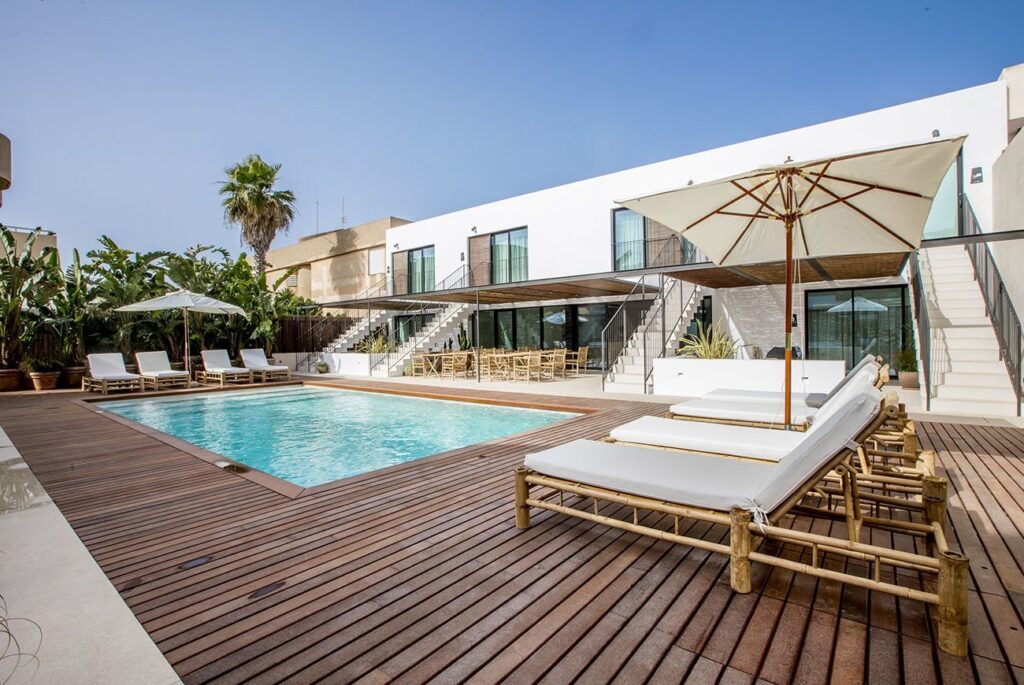 Modern outdoor pool area at one of the boutique hotels in Formentera, featuring a wooden deck, white lounge chairs under umbrellas, and two-story white buildings. Tropical plants and palm trees add lush greenery to the sunny, open space.