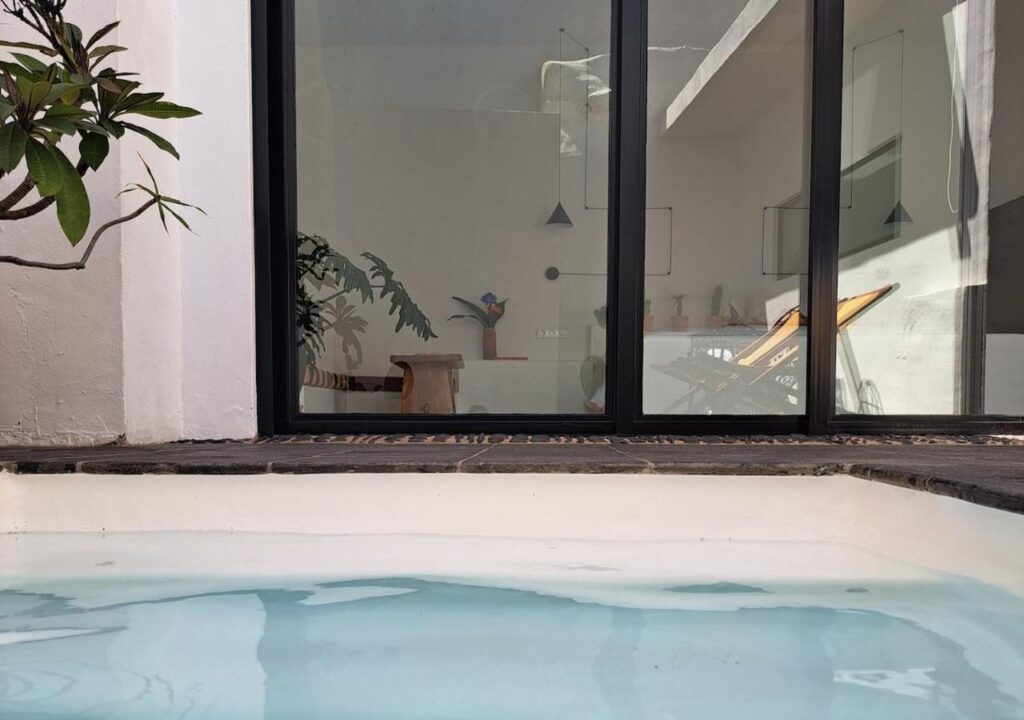 Unique Adults Only Hotels in Lanzarote 7 A view from a small pool at one of the Adults Only Hotels in Lanzarote, looking toward a modern indoor space through large glass doors. Inside are plants, a wooden stool, and a butterfly chair illuminated by sunlight reflecting off the water.