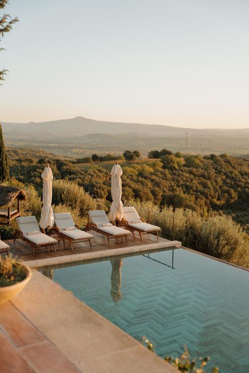 7 Best Agriturismos in Tuscany 18 A serene outdoor pool at one of the finest agriturismos in Tuscany features lounge chairs and closed umbrellas, overlooking a lush green landscape and distant mountains under a clear sunset sky.