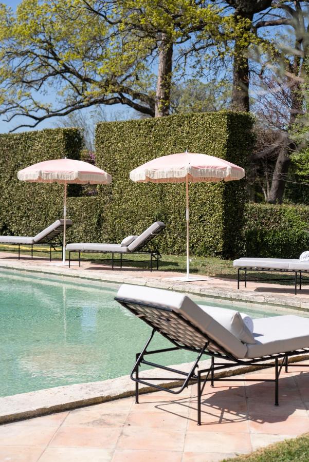 11 Unique Places to Stay in Provence 5 Outdoor swimming pool with clear water, surrounded by lounge chairs and pink umbrellas with fringe. Green manicured hedges and tall trees are in the background on a sunny day—one of the truly unique places to stay in Provence.