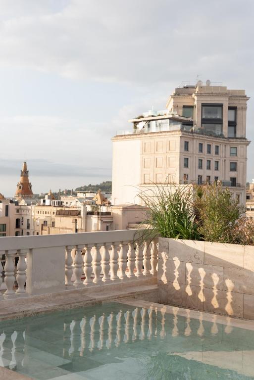 Top Barcelona Hotels with Rooftop Pool 46 A rooftop pool with clear water and stone railings overlooks a cityscape featuring historic buildings and a distant, hazy hillside under a cloudy sky—one of the highlights found at many Barcelona Hotels with Rooftop Pool.