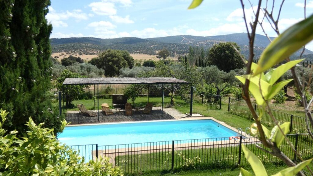 25 Weekend Trips from Madrid Max. 2h Away 11 A fenced outdoor swimming pool is surrounded by green grass and trees, with a shaded seating area nearby—an ideal spot for weekend trips from Madrid. Hills and fields stretch out in the background under a partly cloudy sky.