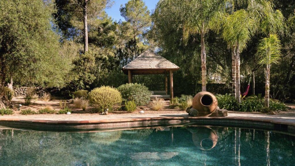 10 AUTHENTIC Finca Hotels in Ibiza 39 A serene backyard evokes the charm of finca hotels in Ibiza, featuring a clear swimming pool, lush greenery, a rustic gazebo with a thatched roof, an amphora pottery piece, and a pink hammock hanging between palm trees.