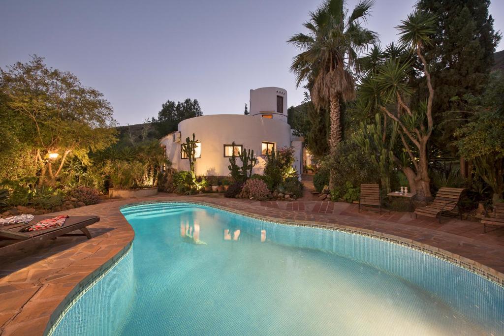 Adults-Only Hotels in Costa del Sol 36 A curved swimming pool with clear blue water sits in front of a white, round stucco house surrounded by lush palm trees—an inviting retreat reminiscent of adult only hotels in Costa del Sol— with lounge chairs on the stone patio at dusk.