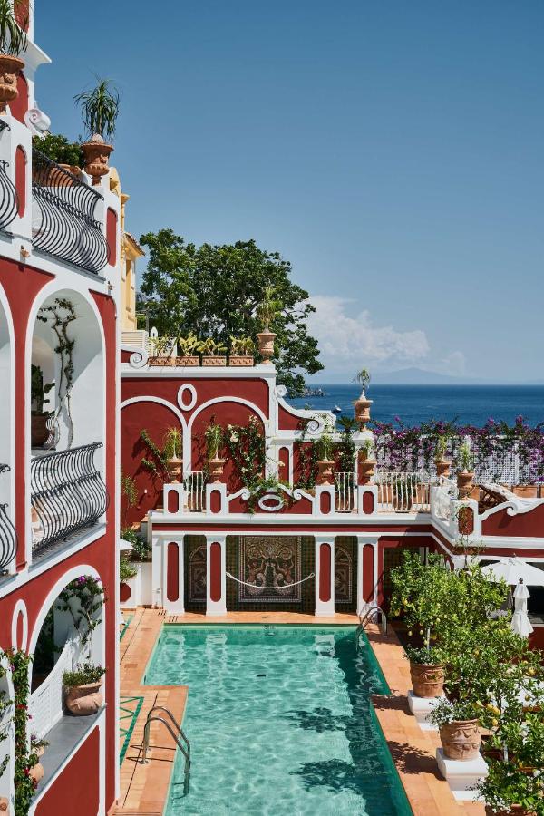 Boutique Hotels on the Amalfi Coast 3 Luxury villa with red-and-white walls, arched balconies, potted plants, and a turquoise pool. Reminiscent of boutique hotels on the Amalfi Coast, it offers Mediterranean Sea views and a serene, sunlit atmosphere under clear blue skies.