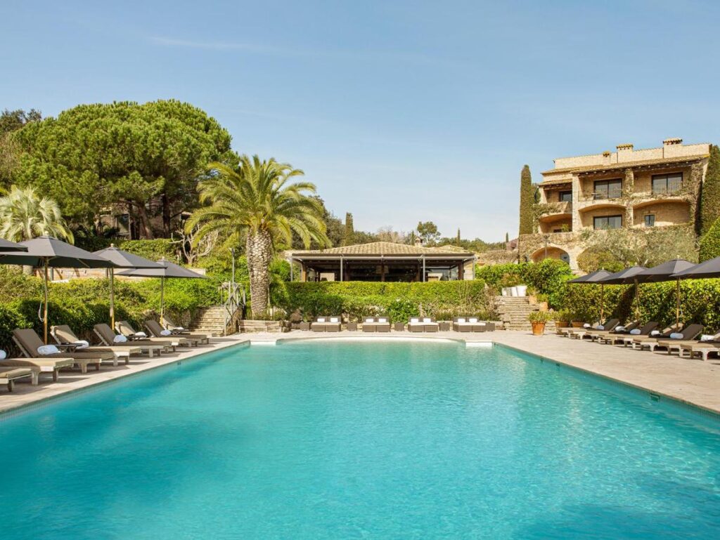 A large outdoor swimming pool surrounded by lounge chairs and umbrellas, with lush greenery, palm trees, and a Mediterranean-style building in the background under a clear blue sky—perfect for boutique hotels in Costa Brava.
