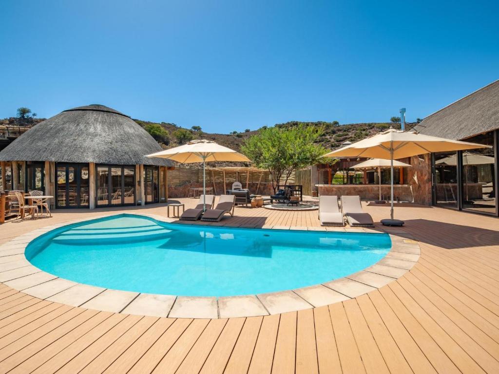 A round outdoor swimming pool with lounge chairs and umbrellas on a wooden deck, surrounded by thatched-roof buildings under a clear blue sky—an idyllic scene often found at Game Lodges in Western Cape.