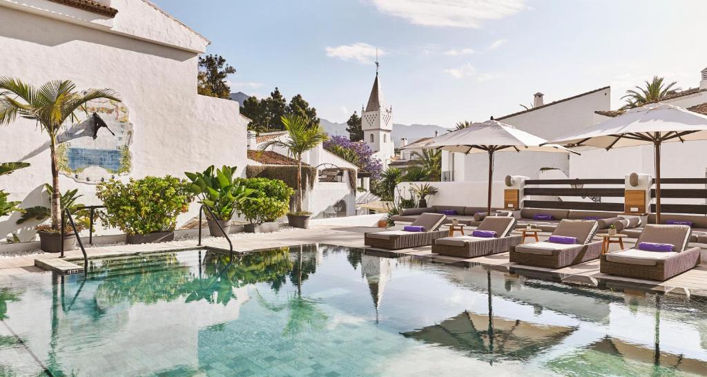 Adults-Only Hotels in Costa del Sol 15 A serene outdoor pool area at one of the top adult only hotels in Costa del Sol features wicker lounge chairs, purple towels, umbrellas, and lush greenery, surrounded by white buildings with views of a church steeple and distant mountains.