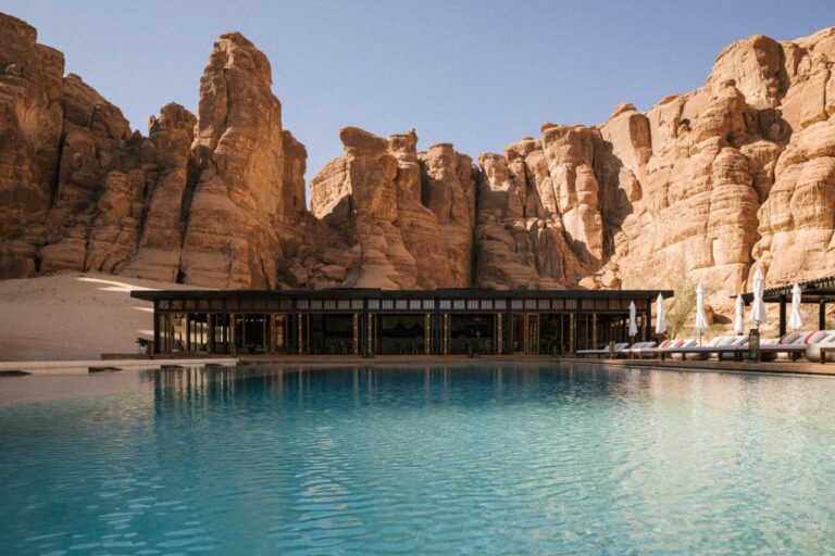 A large outdoor swimming pool with clear blue water is in front of a modern building, surrounded by tall, rugged sandstone cliffs under a clear blue sky—an inviting scene found at some of the best hotels in Saudi Arabia. Lounge chairs and umbrellas line the poolside.