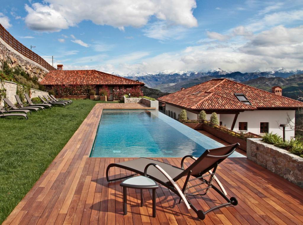 A wooden deck with a lounge chair and small table overlooks a rectangular swimming pool, typical of boutique hotels in Asturias. The pool is framed by grass, stone buildings with red-tiled roofs, and distant mountains under a partly cloudy sky.
