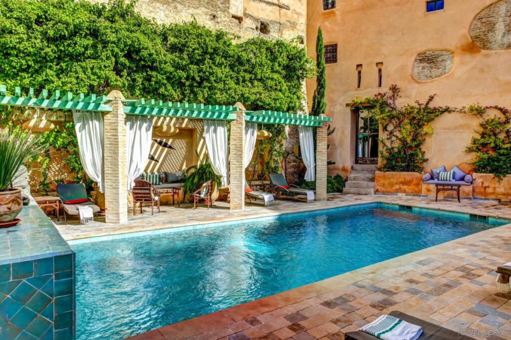 7 UNIQUE Riads in Fez You Won't Forget 26 A Mediterranean-style courtyard reminiscent of riads in Fez features a turquoise swimming pool, tiled deck, shaded lounge chairs under a pergola with white curtains, lush greenery, and rustic stucco walls with climbing plants.