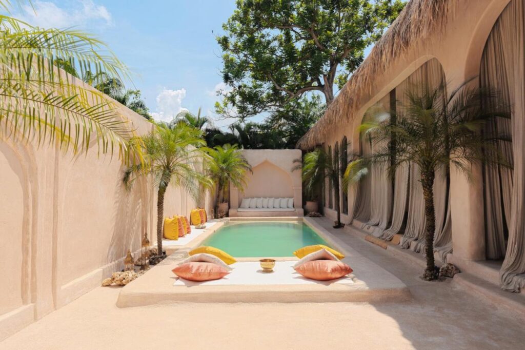 A serene courtyard at one of the best family villas in Bali features a rectangular pool surrounded by palm trees, beige walls, lounge cushions, and sheer curtains. The sunlit space offers a tropical, relaxing vibe with cozy seating and natural decor.