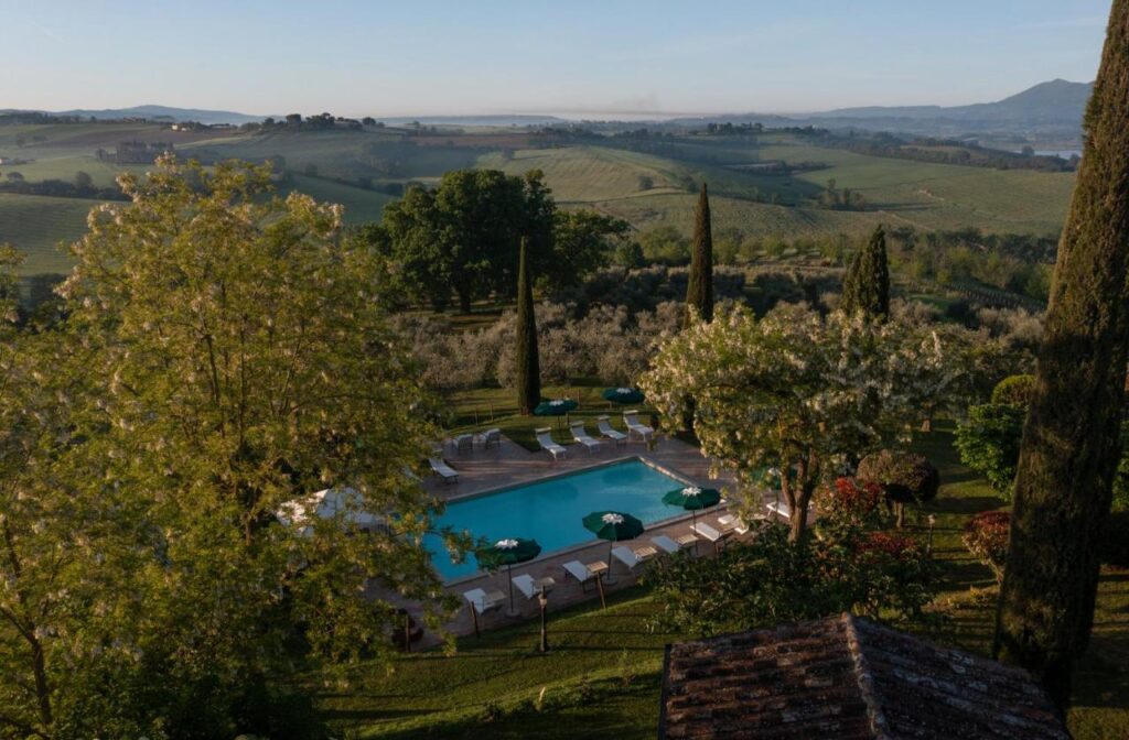 7 Best Agriturismos in Tuscany 3 A serene outdoor pool at one of the best agriturismos in Tuscany is surrounded by lounge chairs and sun umbrellas amid lush greenery, tall cypress trees, blooming shrubs, and rolling hills with distant farmland under a clear sky.