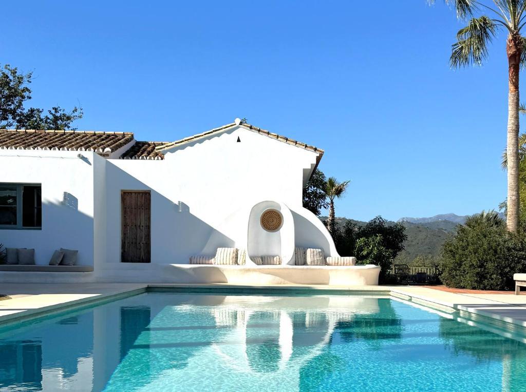 Adults-Only Hotels in Costa del Sol 10 A white Mediterranean-style house with a wooden door and arched seating area is reflected in a clear blue swimming pool—an oasis reminiscent of adult only hotels in Costa del Sol, framed by palm trees, mountains, and a bright blue sky.