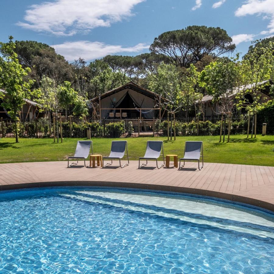 A curved outdoor swimming pool with four lounge chairs and small tables on a wooden deck, surrounded by lush green grass, trees, and tents—offering the relaxing charm found at boutique hotels in Costa Brava under a partly cloudy sky.