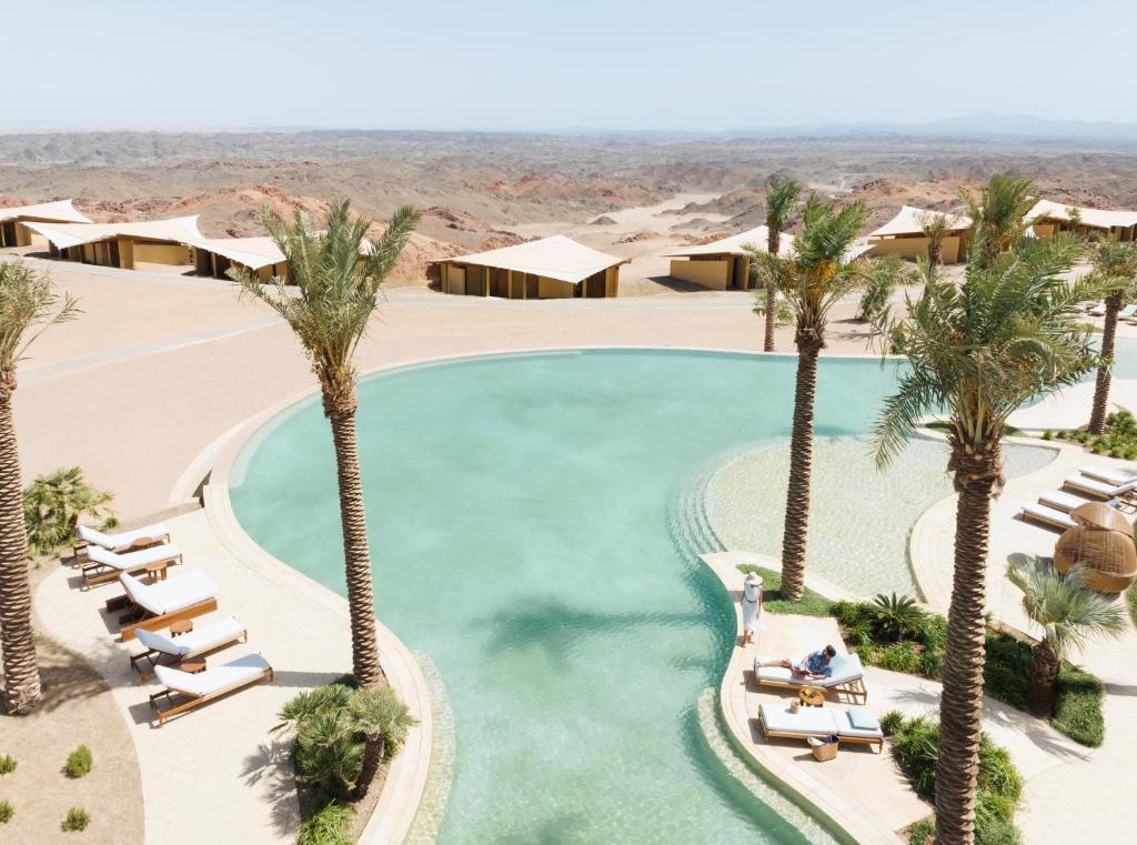 9 Best Hotels in Saudi Arabia: Unique Stays Across the Country 29 A resort pool surrounded by palm trees and lounge chairs sits in a desert landscape, with modern tents and sandy hills in the background—an oasis among the Best Hotels in Saudi Arabia.