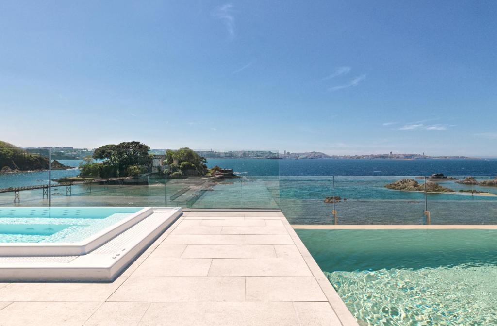 Infinity pool and hot tub overlook a scenic ocean view with glass barriers, a small tree-covered island, and a distant coastal city—an experience typical of boutique hotels in Galicia under a clear blue sky.
