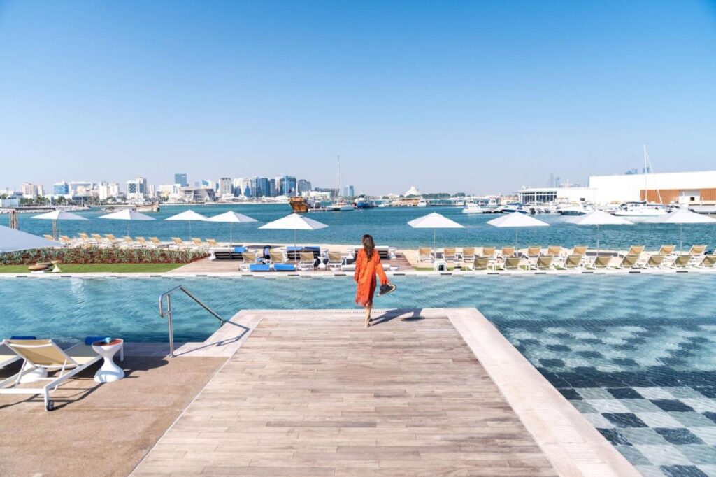 Where to Stay in Doha, Qatar: Transit, City & Beach Hotels 24 A woman in an orange dress walks toward a pool overlooking a marina with yachts and a city skyline—an inspiring scene for anyone seeking where to stay in Doha; rows of lounge chairs and umbrellas line the poolside under clear blue skies.