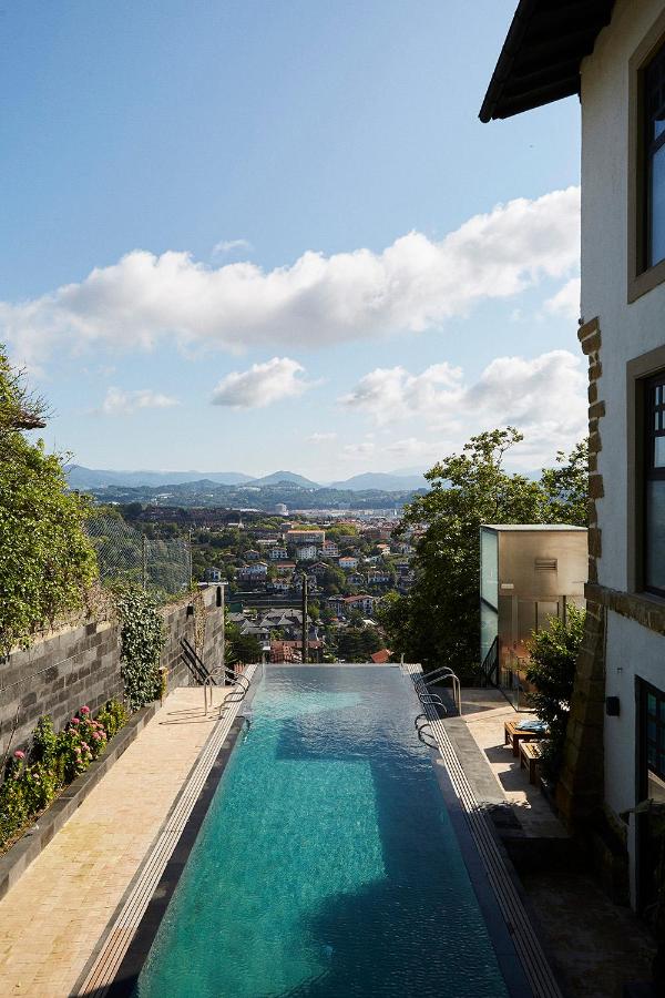 Best Boutique Hotels in San Sebastian 23 A narrow infinity pool at one of the boutique hotels in San Sebastian overlooks a cityscape with distant mountains under a blue sky. The stone patio is bordered by lush greenery and modern buildings.