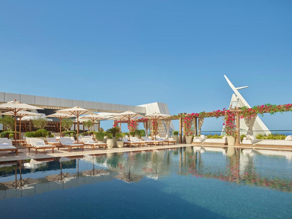 9 Best Hotels in Saudi Arabia: Unique Stays Across the Country 6 A luxurious outdoor pool at one of the Best Hotels in Saudi Arabia, featuring lounge chairs, white umbrellas, potted plants, and floral pergolas beneath a clear blue sky with a modern architectural structure in the background.