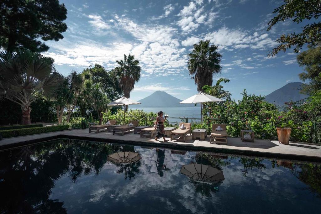 8 UNIQUE Places Where to Stay in Lake Atitlán 2 A woman walks by a pool with lounge chairs and umbrellas, surrounded by lush greenery—a serene scene that hints at where to stay in Lake Atitlan, with views of the large lake and mountains under a partly cloudy sky.