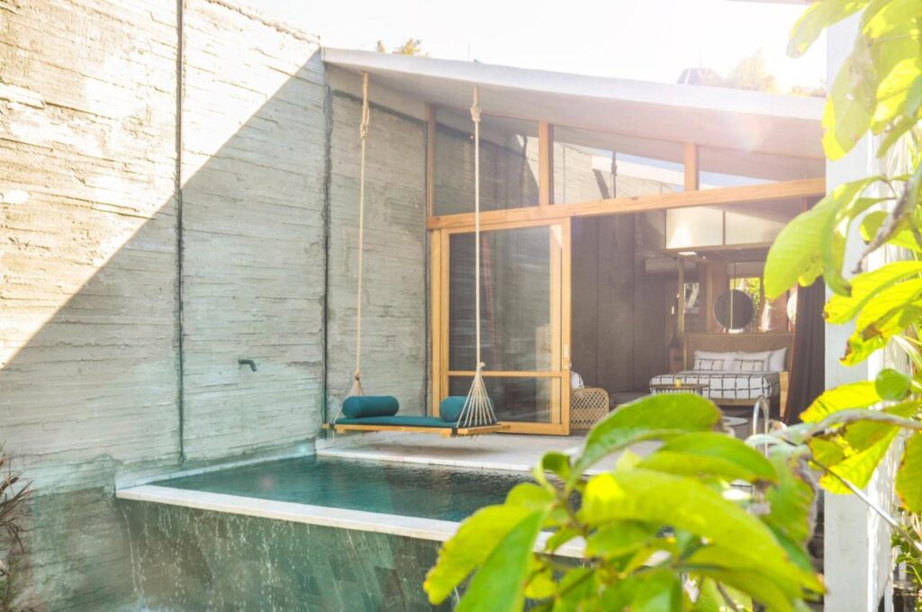 14 UNIQUE Boutique Hotels in Canggu, Bali 19 A sunlit modern villa, reminiscent of boutique hotels in Canggu, features a plunge pool, a wooden swing with blue cushions above the water, large glass doors, and a cozy bedroom inside. Green leaves frame the foreground.