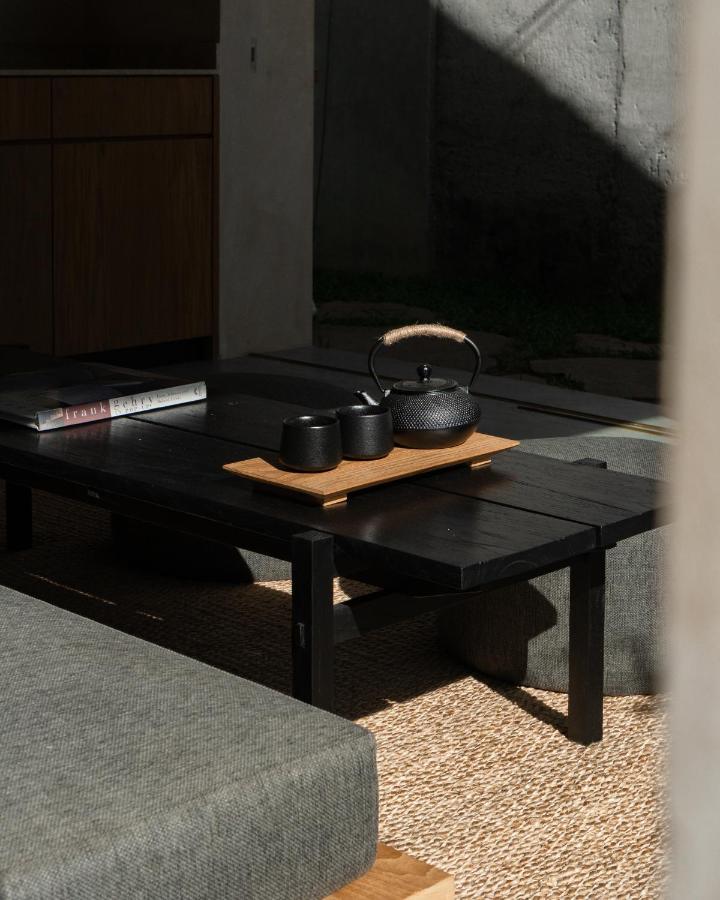 A black teapot and two cups sit on a wooden tray atop a dark low table, surrounded by gray cushioned benches in a sunlit room—just the kind of serene setting you’d find at the best family villas in Bali.