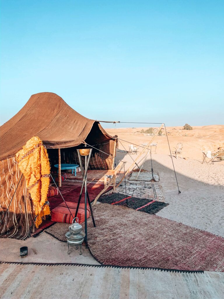 8 SPECTACULAR Agafay Desert Camps 6 A traditional tent with colorful rugs and cushions sits on sandy ground in a desert setting under a clear blue sky, evoking the charm of Agafay Desert camps, with scattered chairs and tables visible in the background.