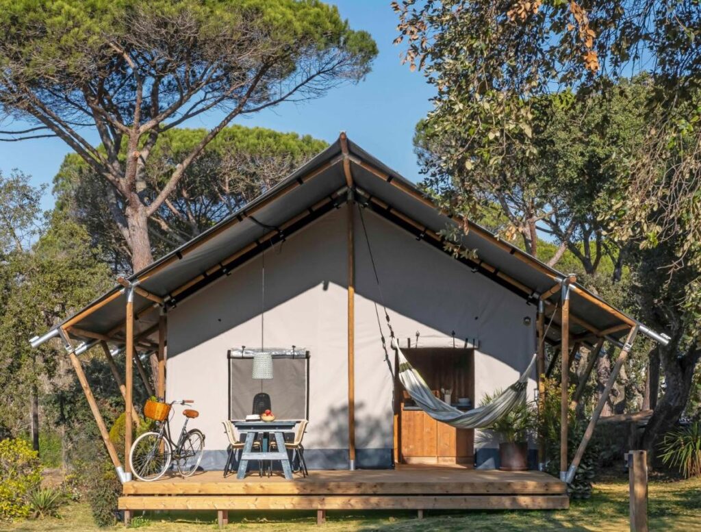A canvas glamping tent sits on a wooden deck in a forest clearing, reminiscent of boutique hotels in Costa Brava. A hammock hangs on the right, a bicycle is parked on the left, and a person works at a table under tall pine trees.