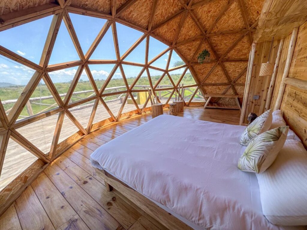 Glamping in the Serengeti: Unique Safari Lodges in Tanzania 3 A cozy wooden dome cabin with a geometric window wall, offering a scenic view of green fields—perfect for glamping in the Serengeti. Inside is a neatly made bed with white linens and patterned pillows, plus wooden chairs on the spacious deck outside.