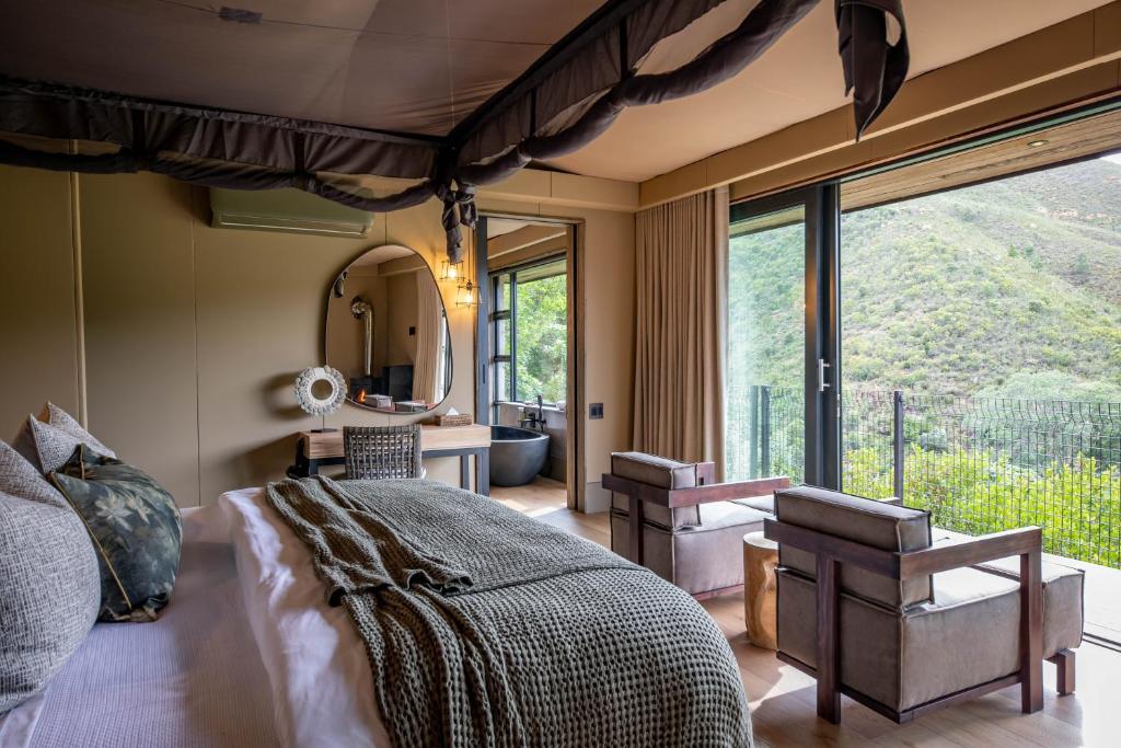 A modern bedroom inspired by Game Lodges in Western Cape, featuring a neatly made bed, textured blanket, two armchairs, a round mirror on a vanity, and large glass doors opening to a balcony with scenic mountain views.
