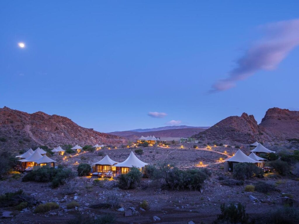 A desert landscape at dusk features several tented Game Lodges in Western Cape with glowing lights, surrounded by rocky hills and sparse vegetation under a clear sky with a visible moon.