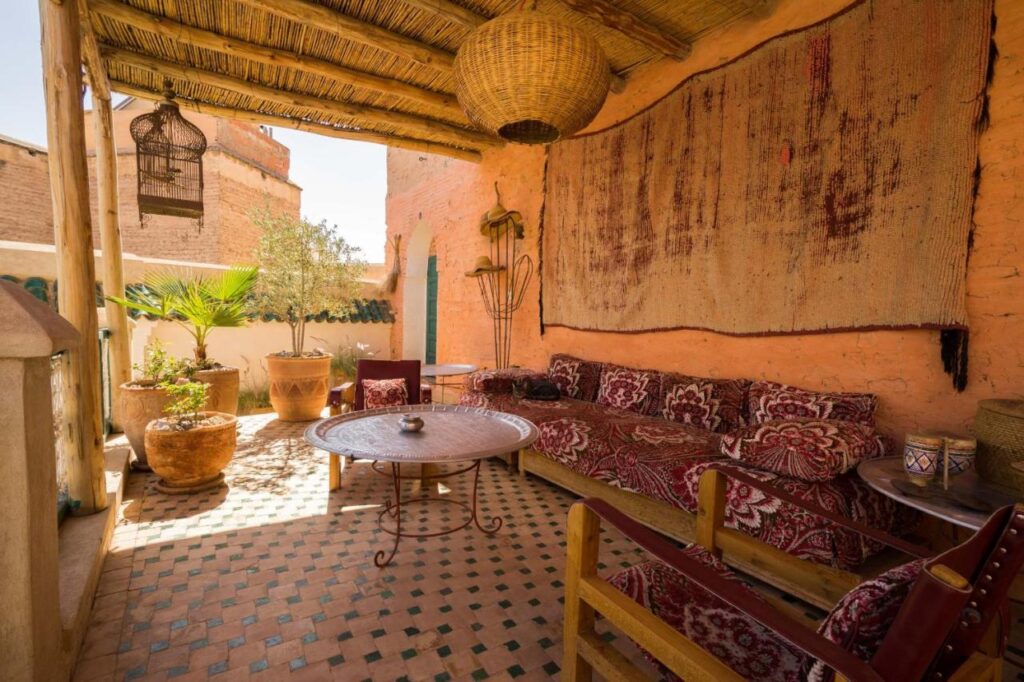 12 Charming Budget Riads in Marrakech 11 A cozy outdoor patio inspired by budget riads in Marrakech, with a wooden roof, patterned sofa and chairs, a round table, potted plants, and a woven rug on a terracotta wall, all in warm earthy tones and rustic decor.