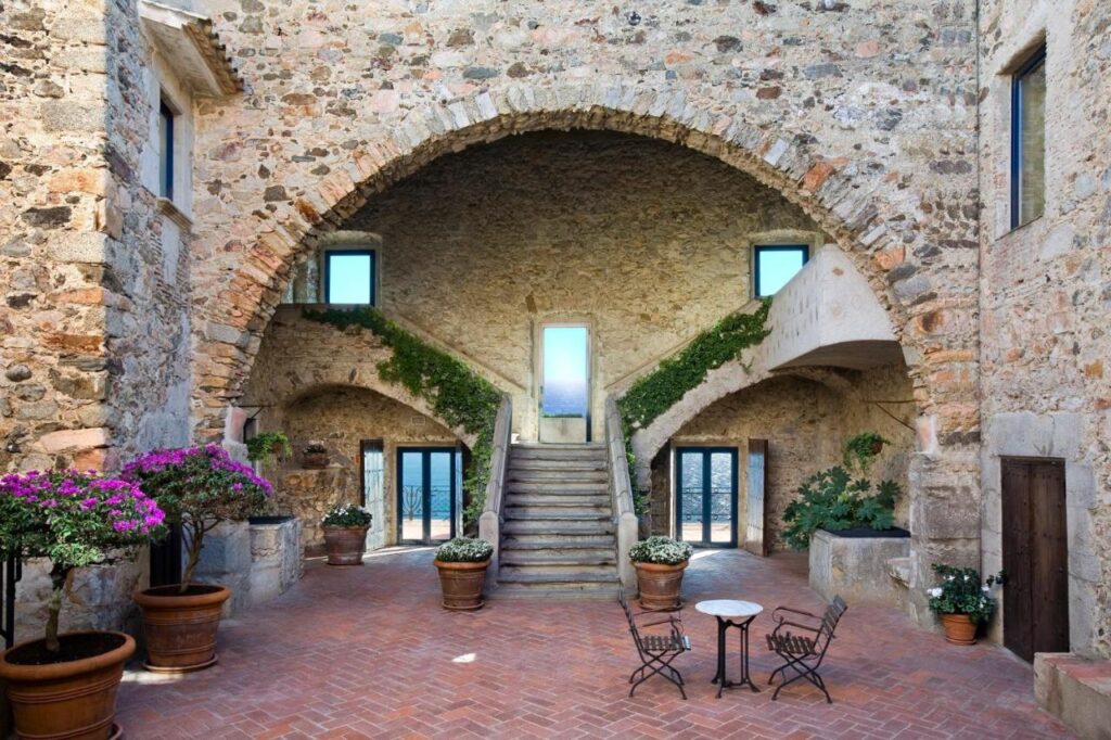 A stone courtyard with an arched ceiling, central staircase, potted plants, and a small table with two chairs on a brick floor—just like the inviting spaces found in boutique hotels in Costa Brava. Large windows and doors open to sea views.