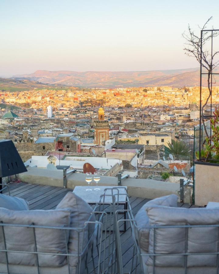 7 UNIQUE Riads in Fez You Won't Forget 16 Two cushioned chairs and a table with glasses overlook a sprawling cityscape at sunset, with distant mountains and a clear sky beyond—evoking the serene ambiance found in riads in Fez, viewed from an inviting outdoor terrace.