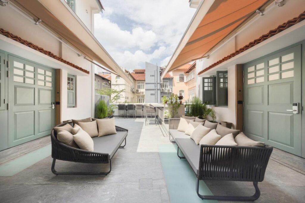 13 Most Unique Hotels in Singapore for an Incredible Holiday 25 Outdoor patio with two modern gray sofas and beige cushions, surrounded by pastel green doors and windows. Retractable orange awnings provide shade, and lush plants add greenery—perfect for guests of unique hotels in Singapore to unwind under the open sky.