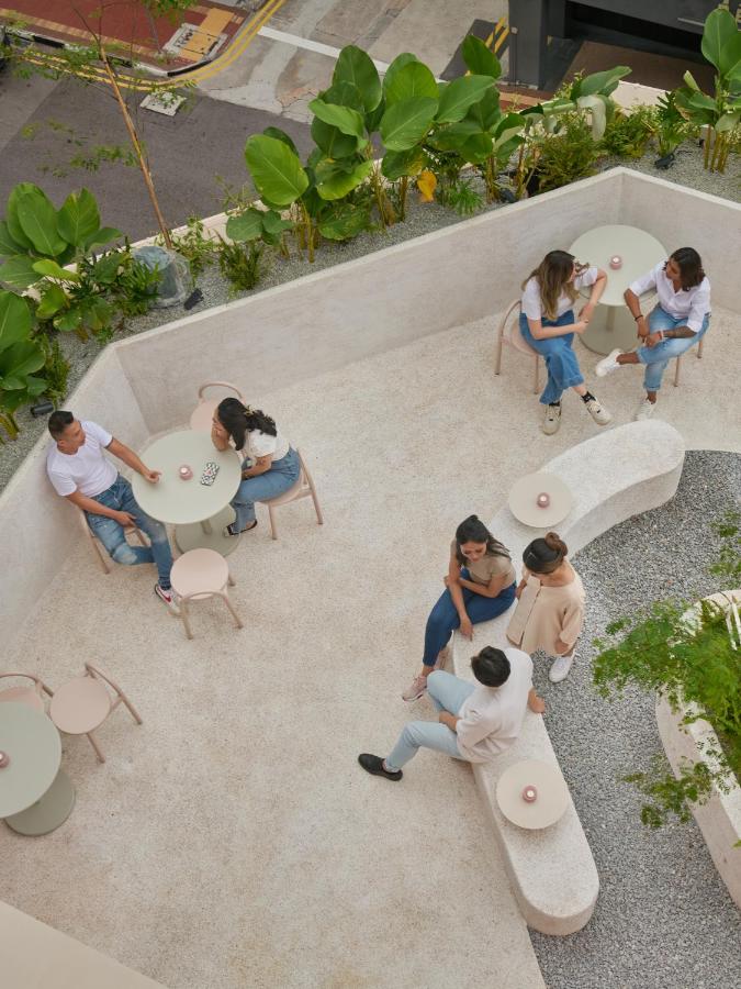 13 Most Unique Hotels in Singapore for an Incredible Holiday 36 A rooftop outdoor seating area at one of the unique hotels in Singapore, with six people chatting in small groups at round tables and benches. The space features light-colored furniture, green plants, and a view of the street below.