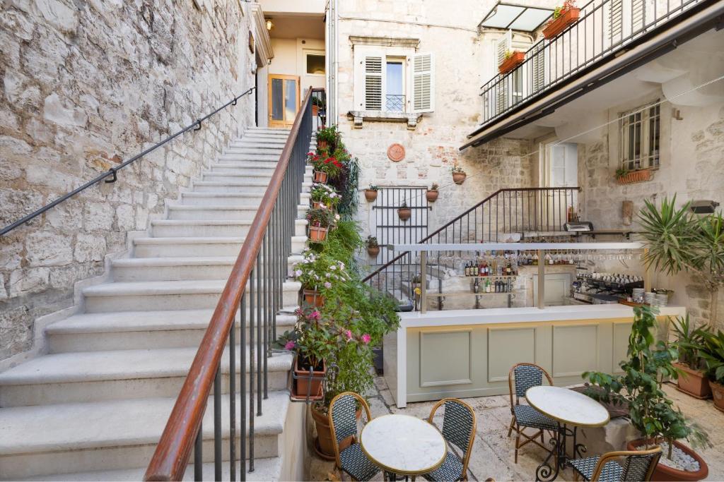 8 TOP Boutique Hotels in Split 4 A charming outdoor courtyard, perfect for those seeking where to stay in Split, with stone walls, a staircase, potted plants, round tables with wicker chairs, and a small bar under a balcony with shuttered windows. Cozy and inviting atmosphere.