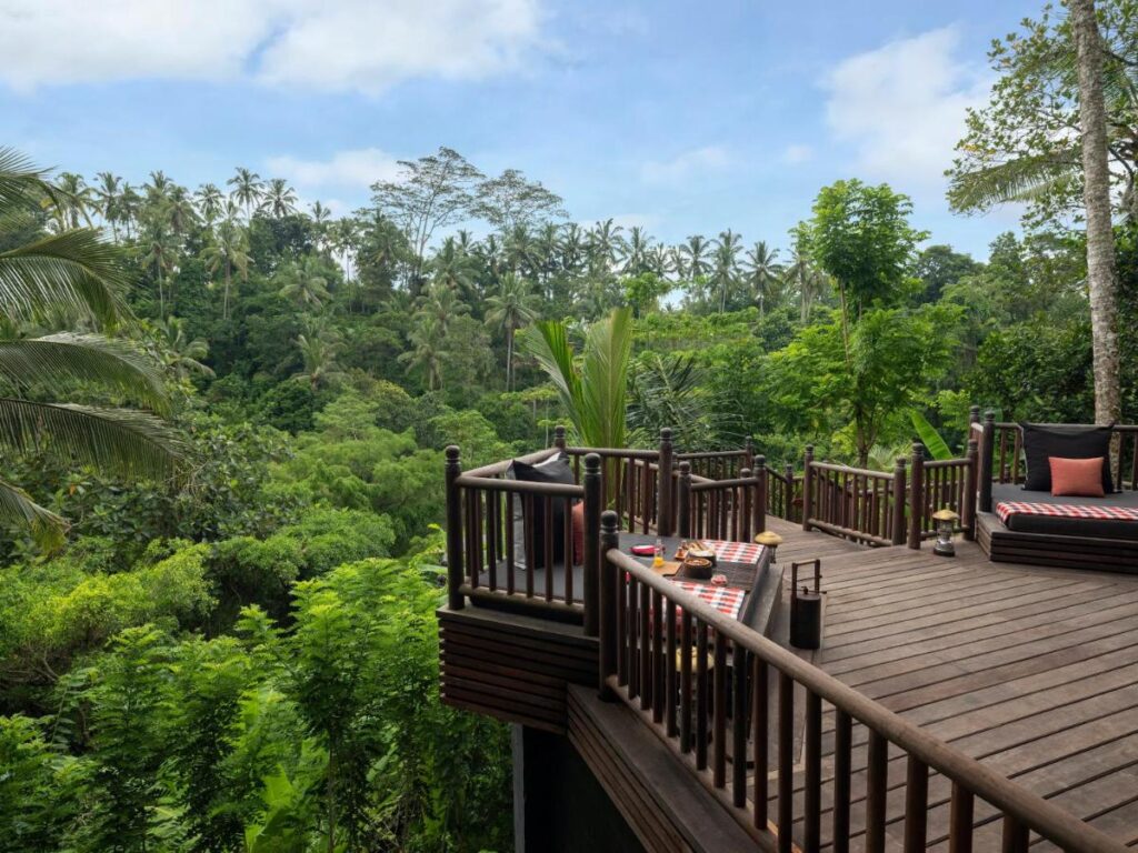 13 Most UNIQUE Places to Stay in Ubud 5 A wooden deck set for dining overlooks lush green tropical forest under a partly cloudy sky, with palm trees and dense foliage—one of the unique places to stay in Ubud for an unforgettable experience.