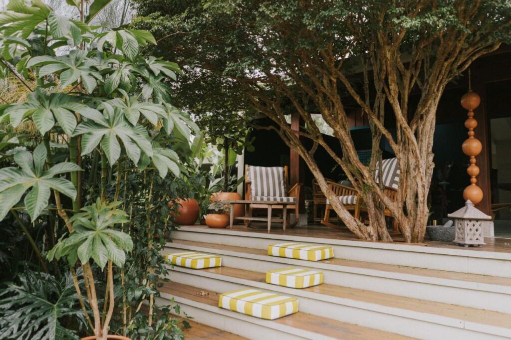 Unique Hotels in Costa Rica: Top Stays 15 A cozy outdoor patio at one of the unique hotels in Costa Rica features striped chairs, wooden furniture, potted plants, and large trees shading white steps adorned with yellow and white striped cushions.
