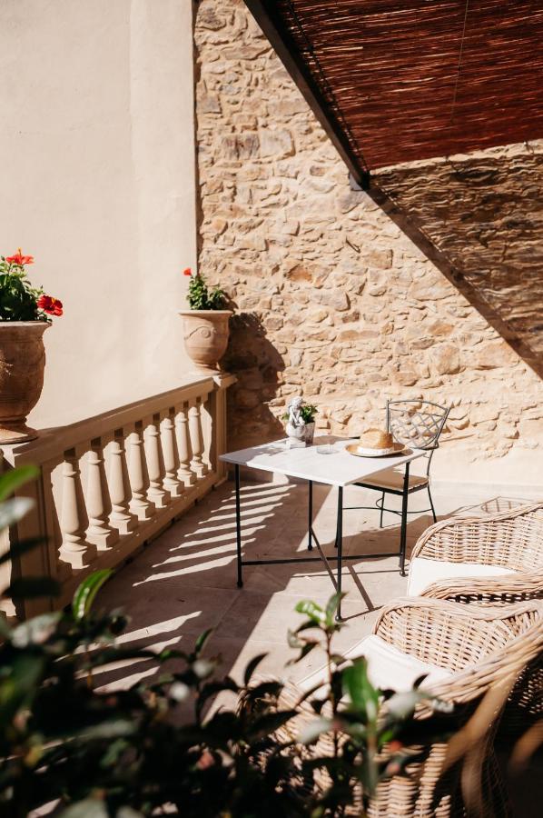 A sunlit patio with wicker chairs, a small table set for two, and potted red flowers against a stone wall—classic charm found in boutique hotels in Costa Brava. Shadows from an overhead cover create a cozy, inviting atmosphere.