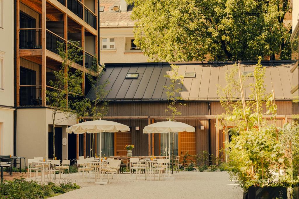 Unique Boutique Hotels in Salzburg 5 Outdoor courtyard with white tables and chairs under large umbrellas, surrounded by green plants—perfect for guests at Boutique Hotels in Salzburg. A modern multi-story building with balconies sits beside a smaller wooden structure, shaded by tall trees.