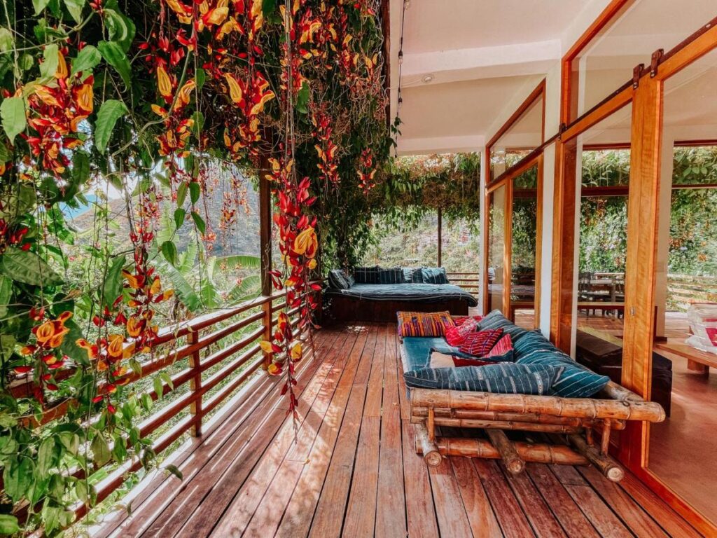 8 UNIQUE Places Where to Stay in Lake Atitlán 12 A cozy wooden patio with bamboo furniture, colorful cushions, and lush hanging plants bursting with red and yellow flowers—perfect for those searching where to stay in Lake Atitlan, right next to large glass windows overlooking the greenery.
